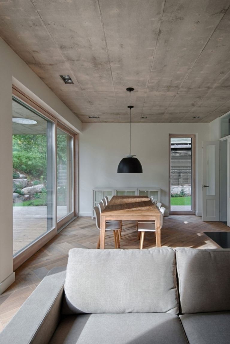 esszimmer-einrichtung-couch-grau-esstisch-stuehle-weiss-haengeleuchte-betondecke-glasfenster-