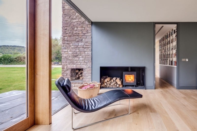landhaus-stil-moderne-architektur-einrichtung-lounge-liege-kaminofen-brennholz-wandfarbe-grau-naturstein-holzboden