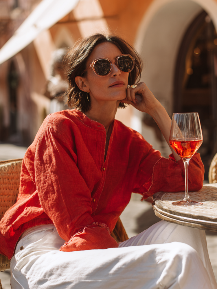 frau mit hemd in beerentönen, weißer hose und sonnenbrille, die wein trinkt