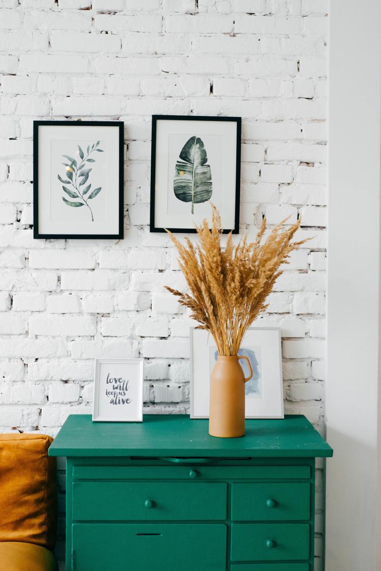 weiß gestrichene Ziegelwand im Wohnzimmer grüne Kommode Bilderwand Pflanzen-Motive schwarze Bilderrahmen