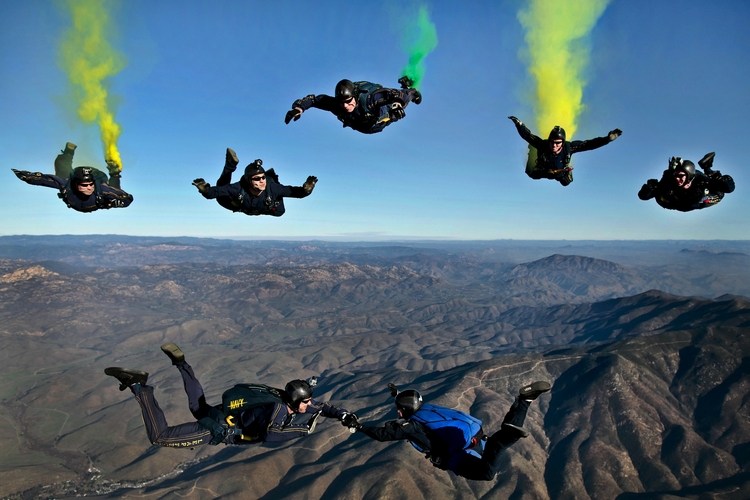 ausgerüstete männer beim fallschirmspringen für junggesellenabschied mit farbigen effekten hoch über berglandschaft