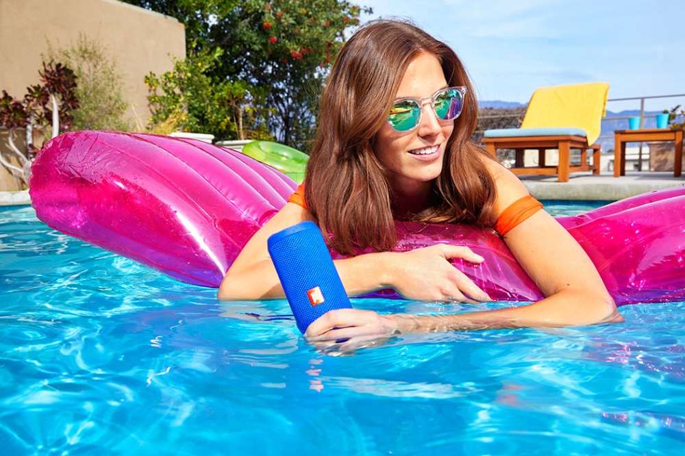 frau mit sonnenbrille liegt unter der sonne im pool auf wassermatratze und hält kabellosen lautsprecher