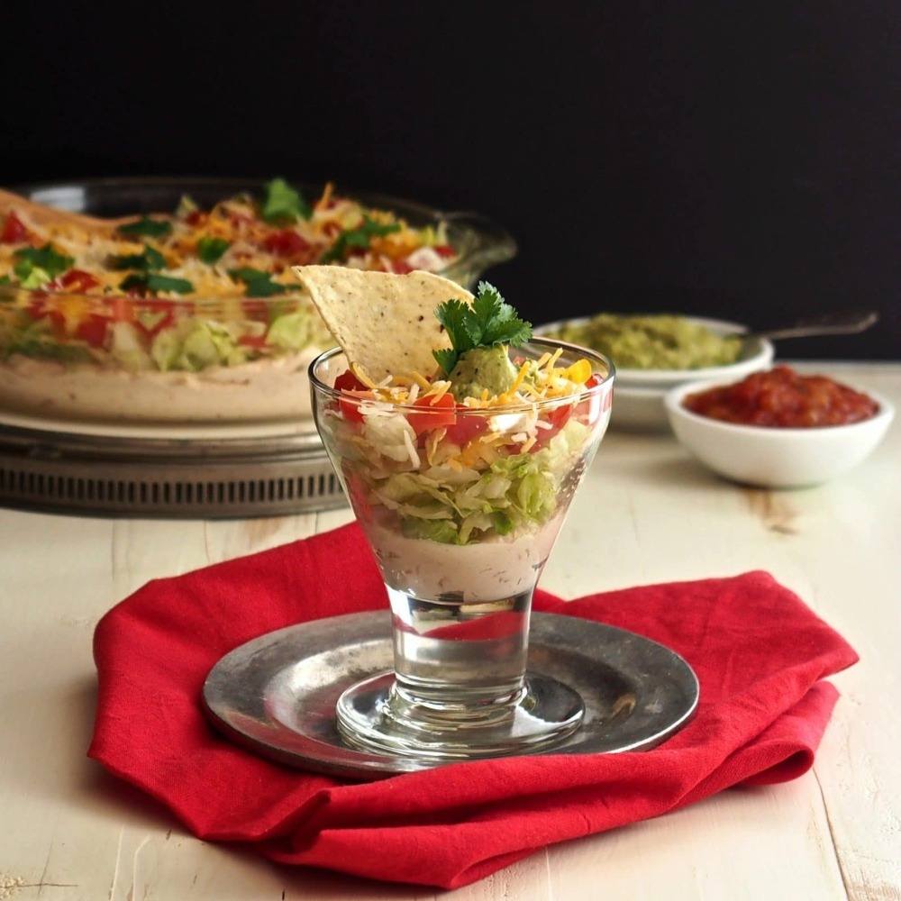 small portion of mexican layer salad in glass on counter with piece of tortilla chips