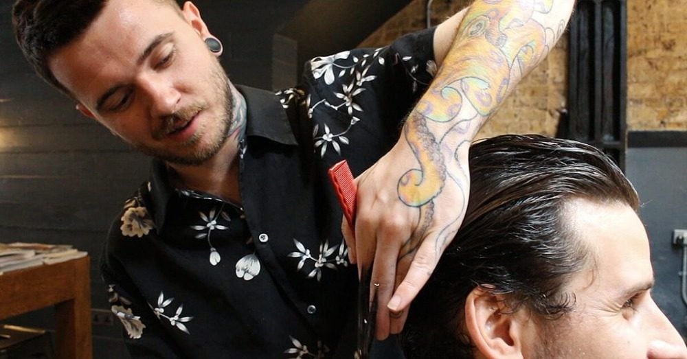 hairdresser with tattoo in arm combs long hair of man in hair dresser