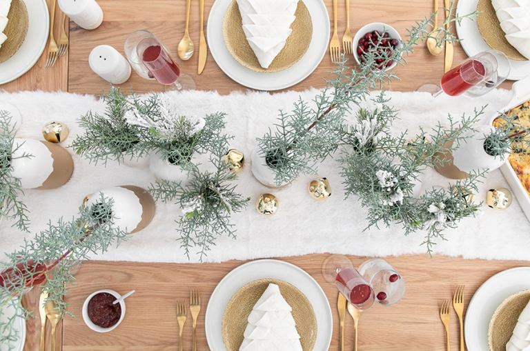 festliche Tischdeko mit immergrünen Baumzweigen und goldenen Keramikvasen und Besteck aus Gold und wie Weihnachtsbaum gefaltete Serviette