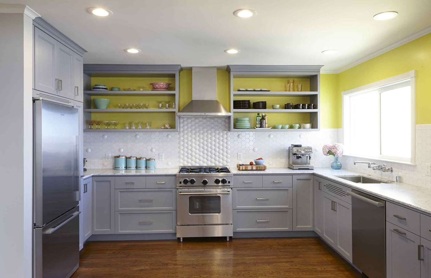 Gelbe Wandfarbe hinter den grauen Regalen und weißer Fliesenspiegel und Edelstahl Küchengeräte