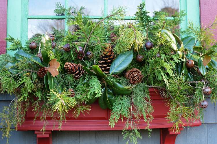 Weihnachtsdeko Fensterbank aussen Tannenzapfen und frische Blätter und Tannengrün und Weihnachtsglocken im Blumenkasten arrangieren