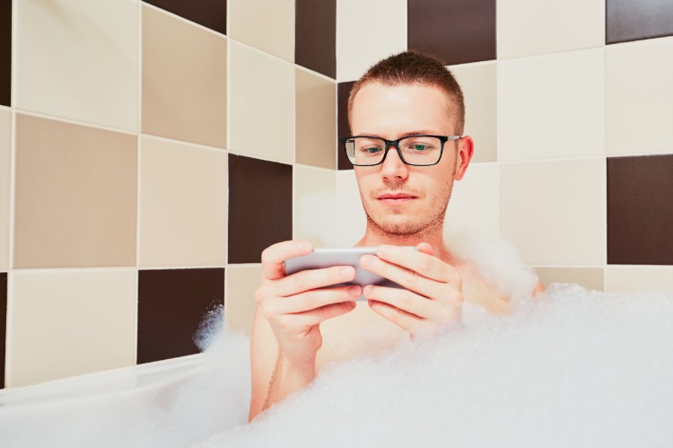 junger blonder mann mit brille nutzt handy in badewanne
