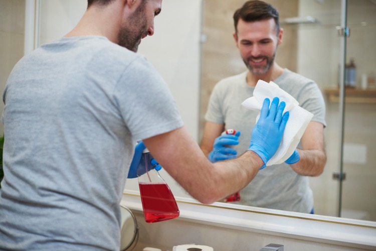 lächelnder mann mit gummihandschuhen putzt badezimmerschrank mit spiegelfassade