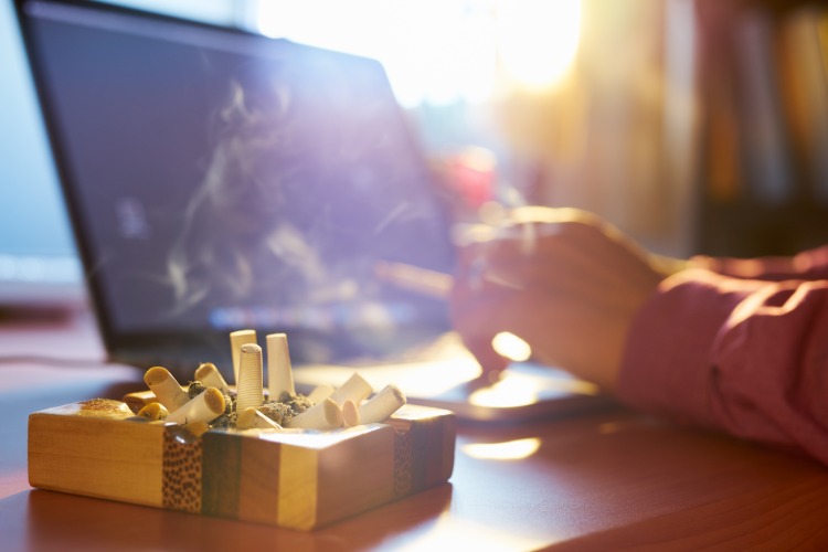 aschenbecher voll mit zigarettenkippen neben rauchender mann vor laptop