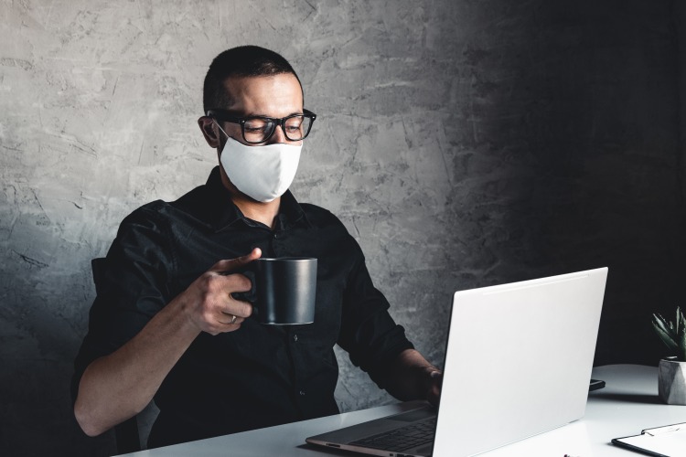 mann mit brille und mundschutz hält kaffeetasse vor laptop