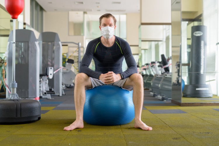 mann mit schutzmaske sitzt auf ball im fitnessraum