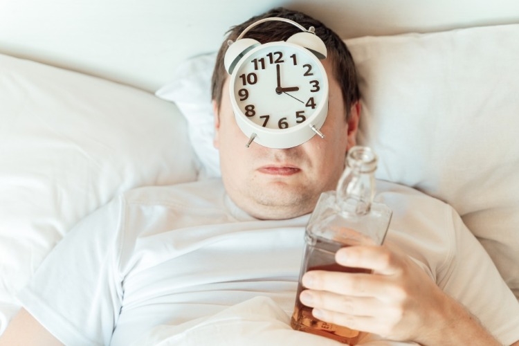 verkaterter mann im bett mit wecker und flasche mit whisky