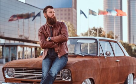 bärtiger mann mit brauner lederjacke im klassischen stil und jeans auf einem verrosteten retro wagen