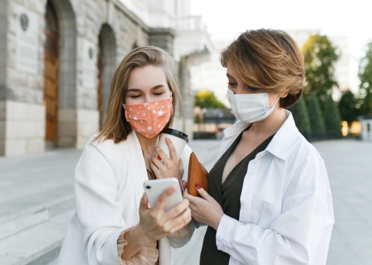 zwei junge frauen mit schutzmasken besprechen östrogen und progesteron bei coronavirus auf dem handy