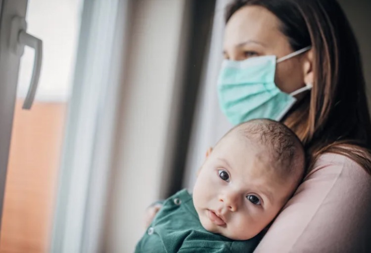 mutter mit schutzmaske gegen coronavirus hält ihr baby vor einem fenster