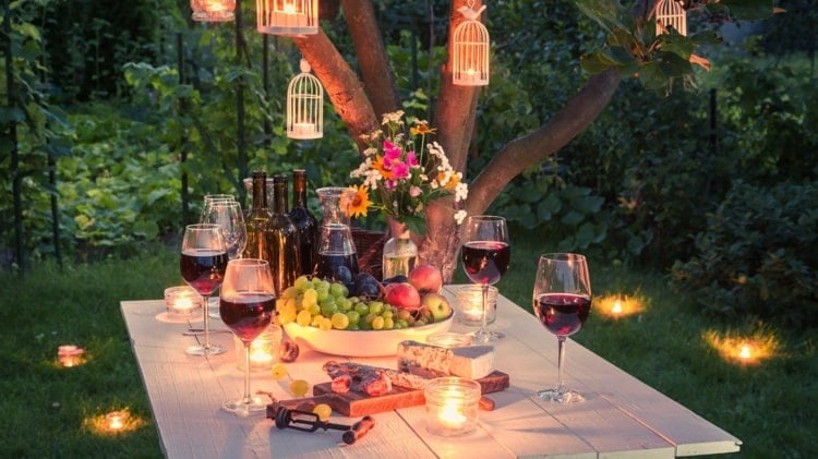 Esstisch mit schöner Gartenparty Beleuchtung - Kerzen auf dem Tisch, im Rasen und hängend am Baum in Vogelkäfigen