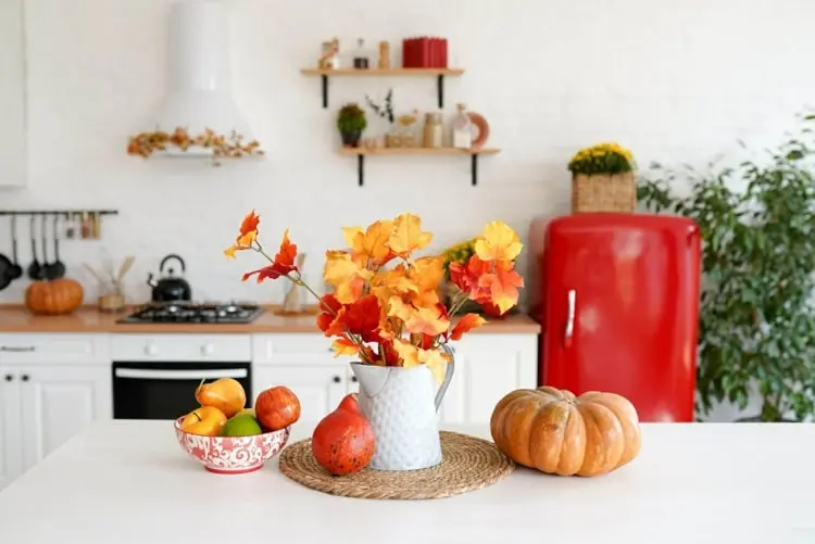 Weiße Kanne mit buntem Herbstlaub als Tischdeko in der Küche