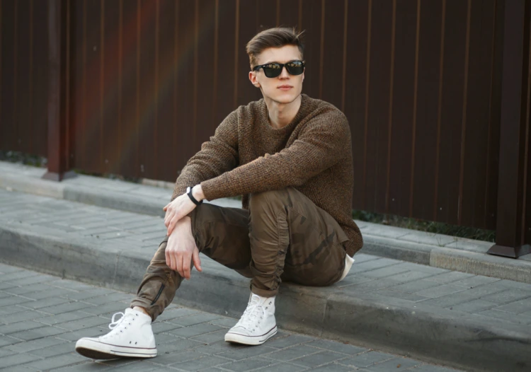 mann mit braunem pullover, camouflage cargo hose und weißen converse sneakern