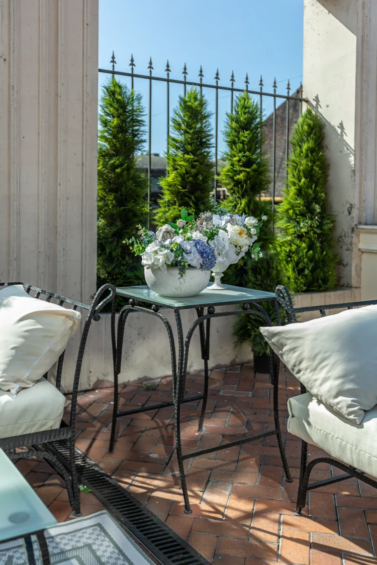 eine schicke terrasse eines landhauses mit schmiedeeisernen möbeln, vasen mit blumen und sträuchern als sichtschutz