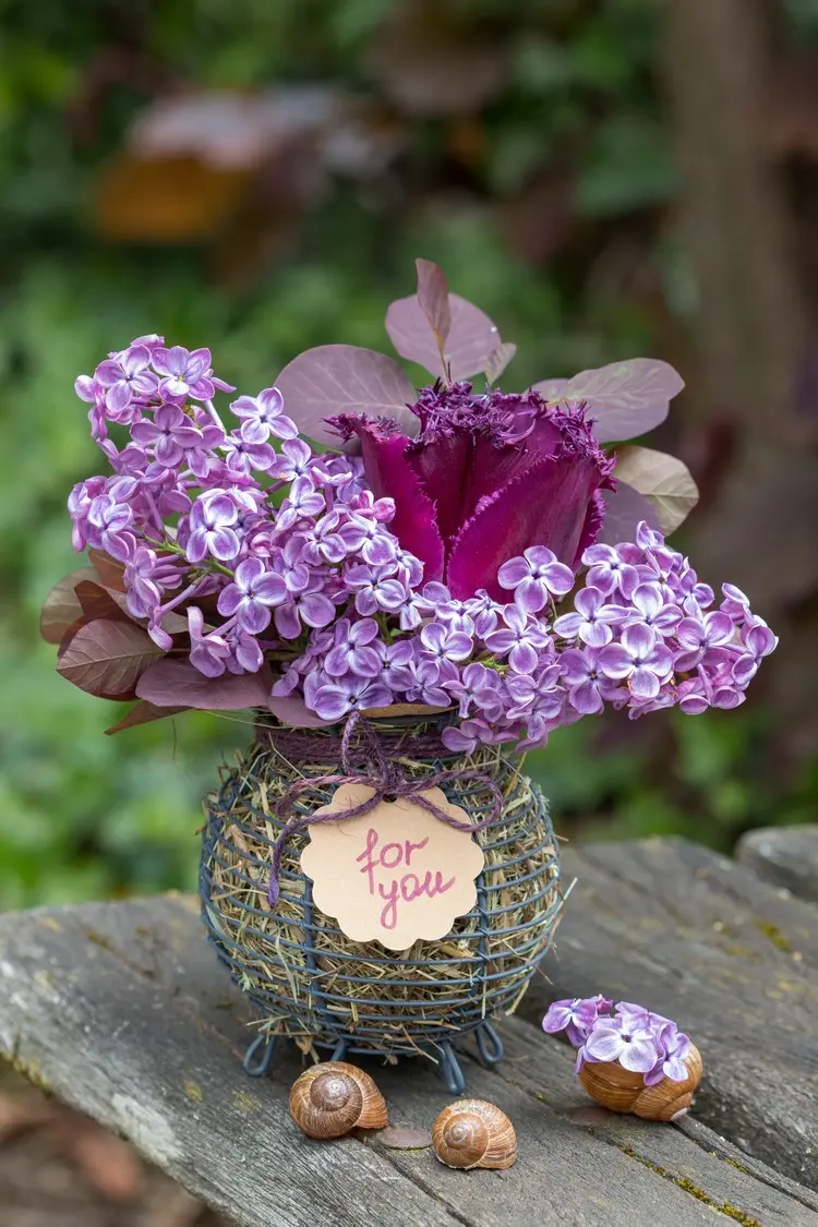 Korbgefäß mit Heu als Vase für Fliederblüten, Papageien-Tulpe und rote Blätter dees Spierstrauchs